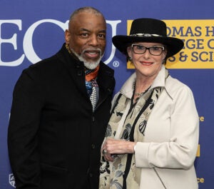 A nearly bald man with a white mustache and goatee, wearing a colorful scarf and black suit jacket, stands next to a woman wearing a black hat, glasses, a seashell scarf and a beige suit jacket. They are in front of a purple and gold East Carolina University Thomas Harriot College of Arts and Sciences photo backdrop.