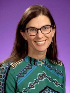 A women in a green patterned shirt, brown hair and glasses sits in front of a purple photo backdrop