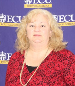 A woman in a red shirt and blond, shoulder-length hair stands in front of a purple banner with a white and gold Political Science department logo.