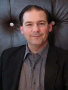A man in a dark suit jacket and gray shirt sits in front of a dark leather background