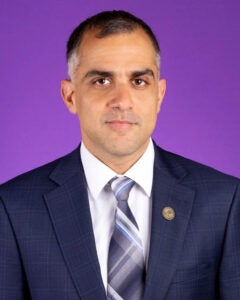 A man in a blue suit jacket and tie sits in front of a purple photo backdrop