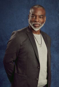 LeVar Burton in a beige sweater, dark blazer, and blue backdrop