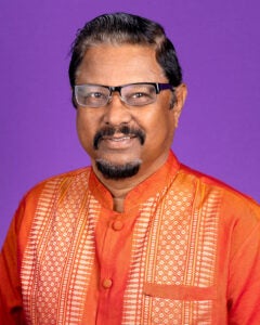 A man in an orange shirt sits in front of a purple photo backdrop